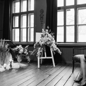 Grayscale funeral scene showing a woman in mourning by a coffin and floral arrangements.
